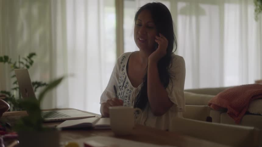 Woman with white shirt and wavy hair sits at table talking on smartphone in the living room at home