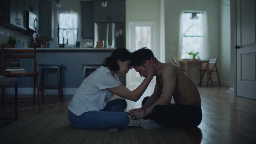 A man and woman sit on the floor facing each other when the woman takes off her necklace and puts it around the man then gets up and walks away