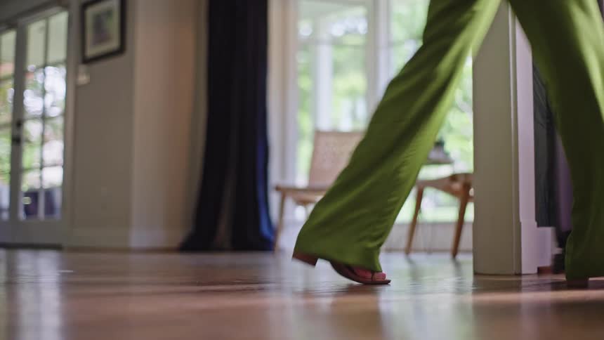 A person in green pants walks across a room to the door while rolling a suitcase