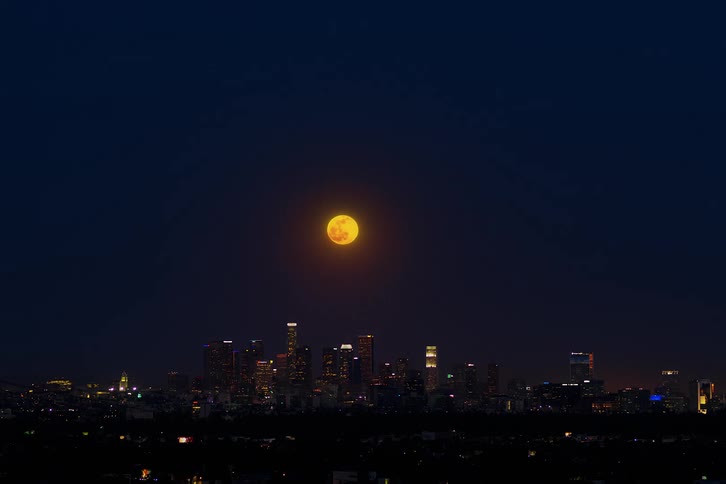 A time lapse of a wide city skyline as the moon and dark sky come up over the buildings