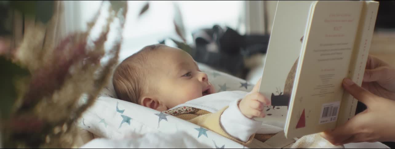 Mother holds picture book in front of baby lying on its back in cradle
