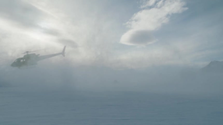 Helicopter takes off leaving person with bags on snow covered plain with mountains in background