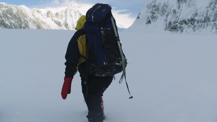 Man with yellow coat red gloves and black pants hikes through snow on the mountain