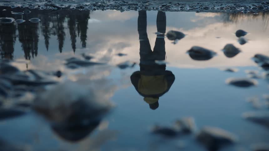 A close up of shallow water reflecting the sky trees and a person walking and then standing