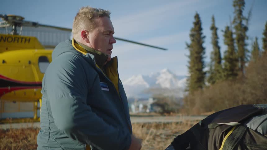 Man puts on cold weather gear while standing outdoors with helicopter nearby and mountains in the background