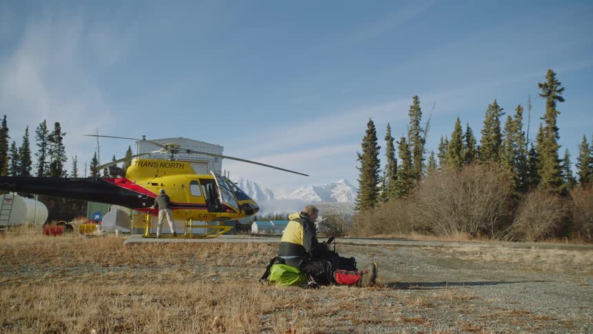 A man sitting on the ground going through his bags while another man moves around a helicopter parked
