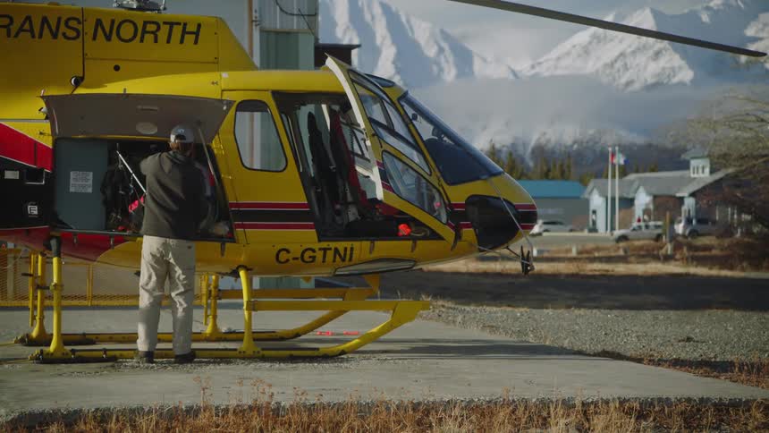 Man unloads bag from helicopter a distance from buildings and mountains then walks away