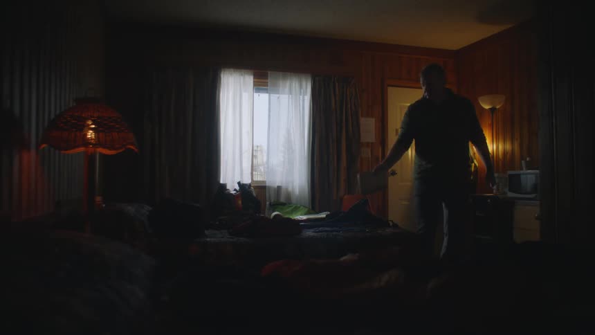 A man is slowly unpacking a bag before sitting down on a bed in a motel room in the evening
