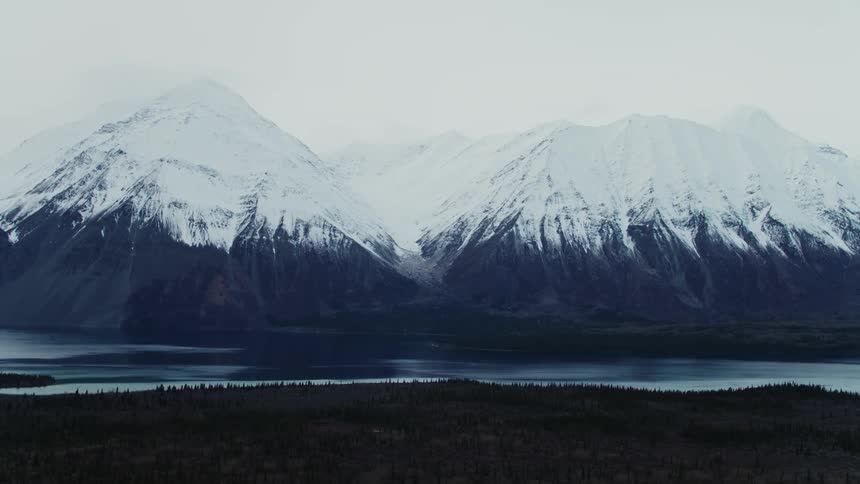 Snow covered mountains reflect from water across lake from forest