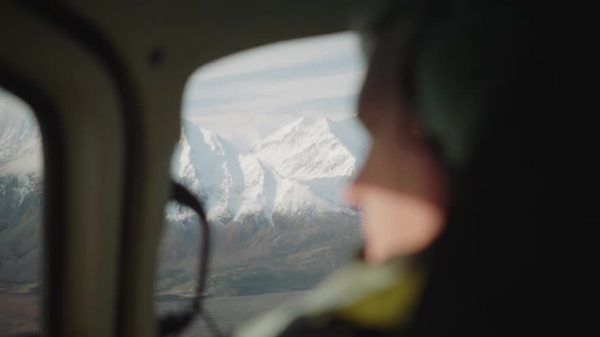 A man piloting a hovering helicopter is staring to the left searching for something in a mountainous region during the daytime