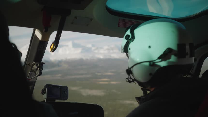 Two pilots in the cockpit are talking as their helicopter flies over land toward snow covered mountains