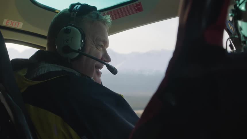 A man is wearing a headset as speaking to the person sitting next to him as he flies a helicopter near snow covered mountains