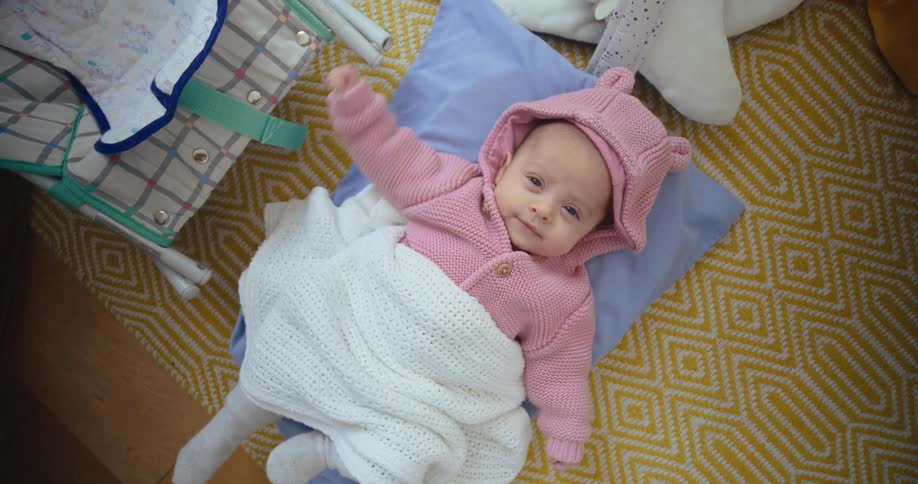 Camera moves towards a baby in pink hoodie lies on yellow patterned rug and kicks her feet while smiling
