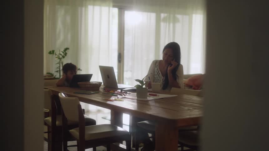 A mother and son sitting at a dining room table the mother on her phone and laptop while the boy sits looking at a tablet