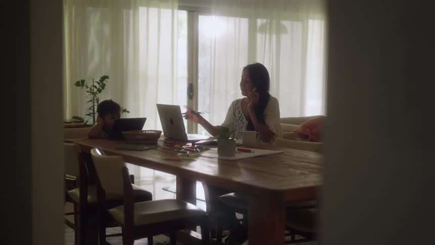 A mother and son sit at a dining table the mother on her cell phone while using her laptop and the boy watching a tablet