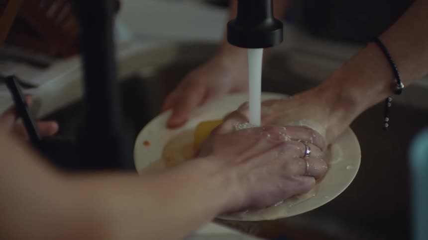 A close up of a persons hands washing dishes when another person puts their hand over the first persons