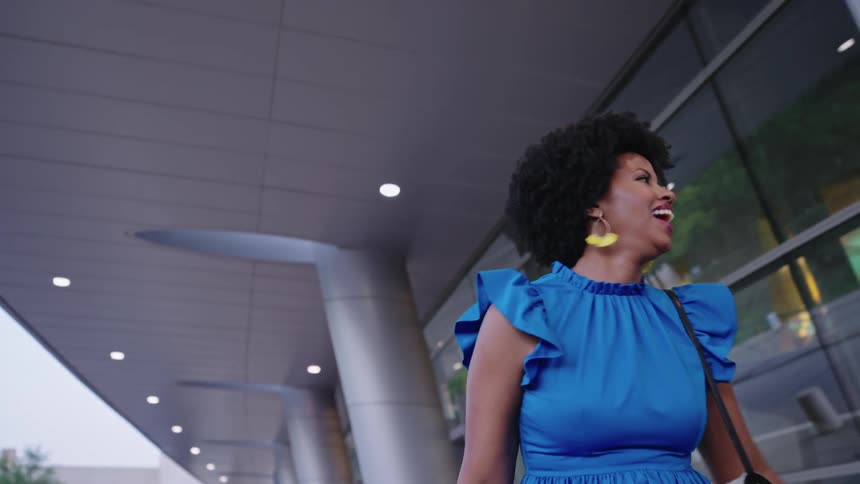 A woman walks alongside a large glass wall moderling a blue blouse and purse while smiling and waving