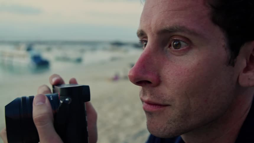 An up close of a man holding a camera up to his face taking a picture then smiling and looking at the camera