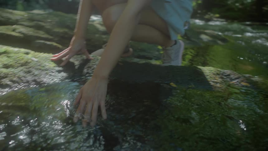 A woman kneeling beside a stream runs her fingers through the waters