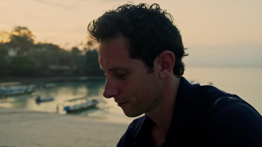 Man sitting on beach texts on smartphone and looks over toward boats moored near beach under a red tinted horizon