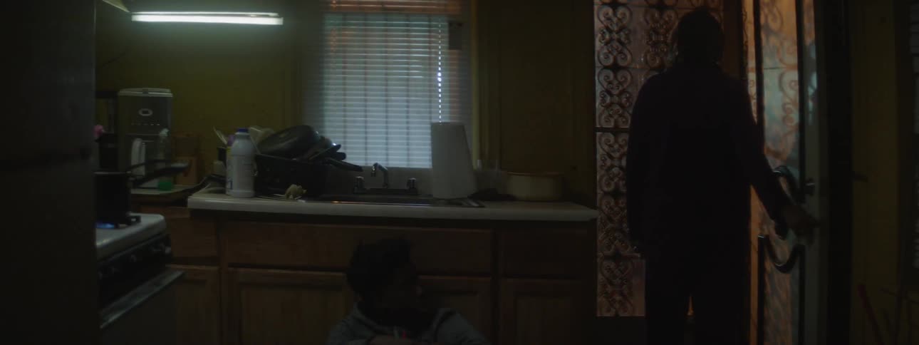 A man looks out his screen door while another man hides behind a cabinet