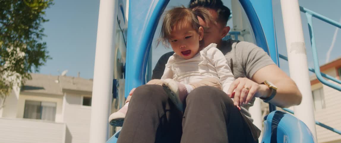 A man puts his baby daughter in his lap as they ride down the slide on the playground and his wife is smiling at them at the end of the slide
