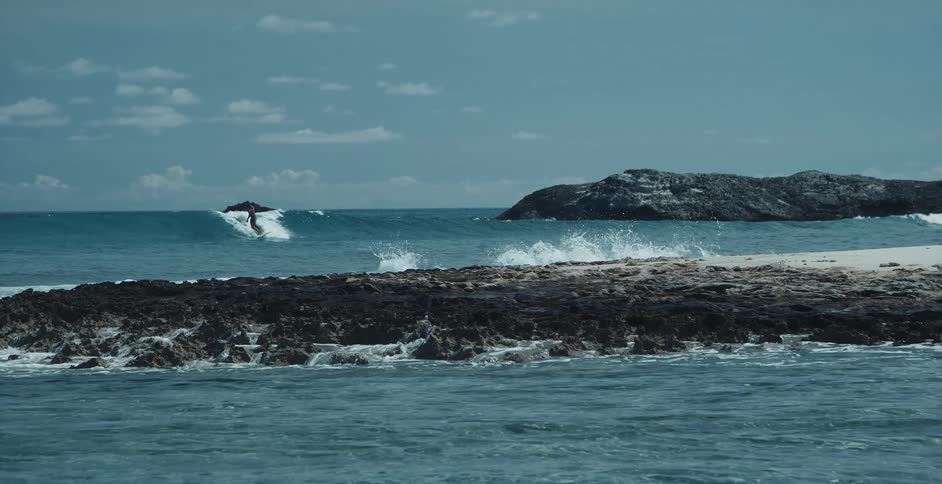 A person surfs toward a beach shoreline among rocky outcroppings on the sea