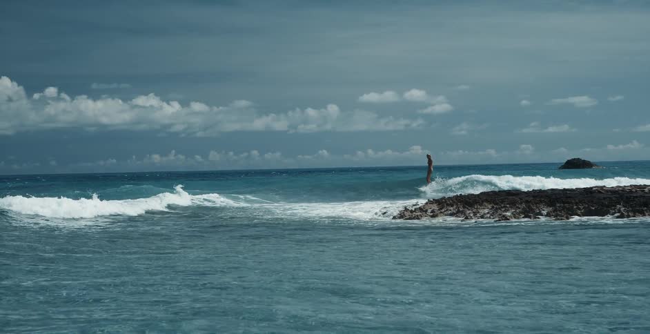 Surfer rides wave near low rocky island before falling