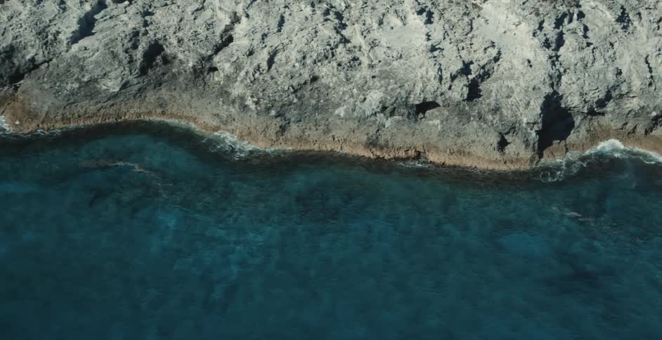 An aerial view of a deep turquoise sea as it meets a grey rocky coastline