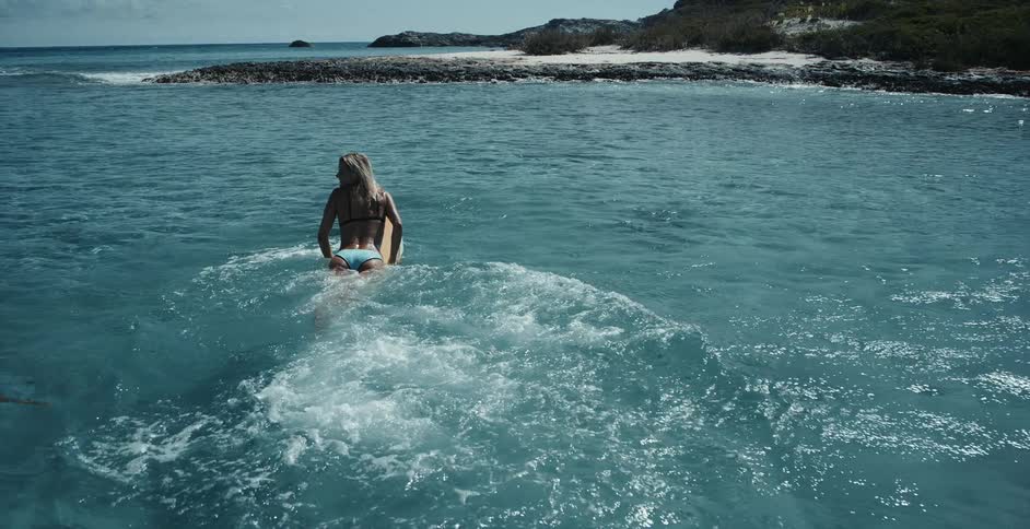 Woman wearing a bikini jumps onto surfboard in the ocean near the shore and begins to paddle out