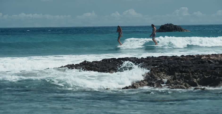A man and woman on surfboards riding a wave to shore near a rocky cove