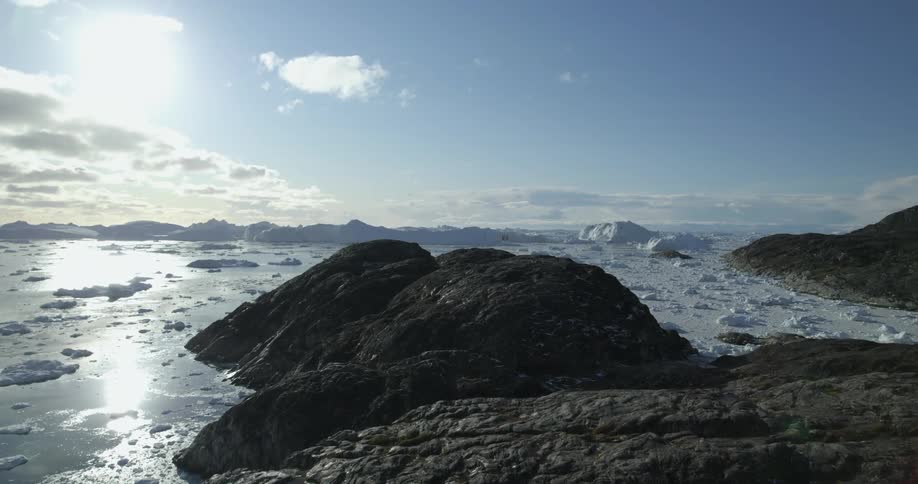 An aerial of coastal rocks in the arctic ocean with icebergs and mountains in the ocean