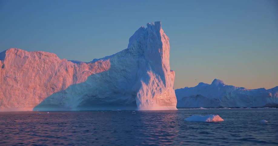 Moving across ocean icy waters past glacier with pink light cast on it from the sunrisesunset