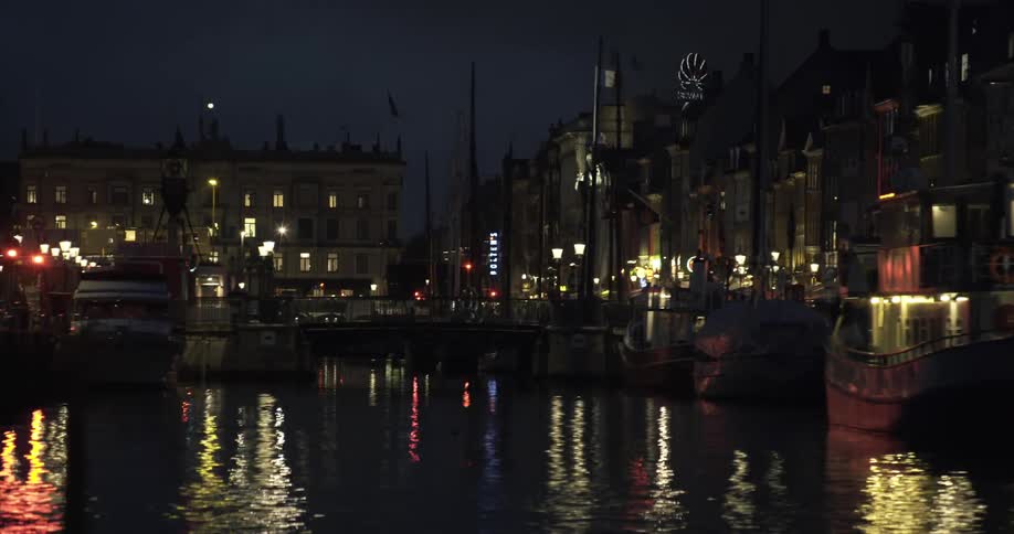 Boats parked in a pier in a small waterway between buildings with lights from the city reflecting off the water