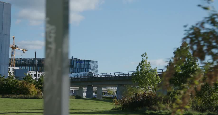 A monorail train moves over a bridge in a grassy urban area toward distant buildings