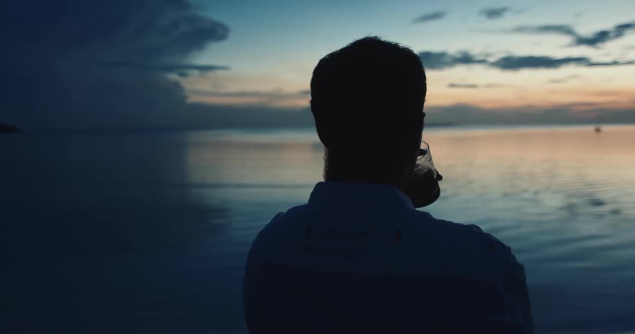 A man is standing along the coast looking at the water and drinking from a glass as lightning flashes in the sky