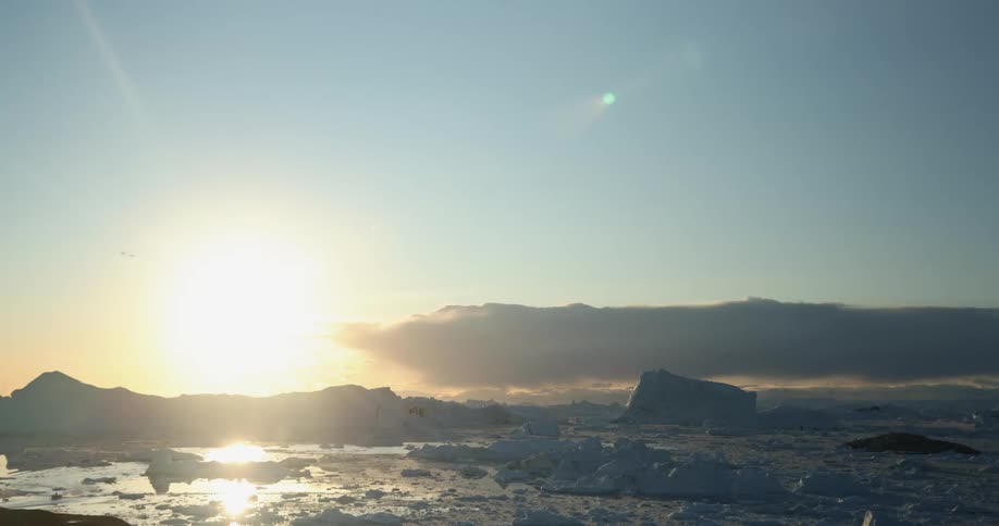 Sun sinks toward horizon and sets briefly glaring across sea ice and icebergs