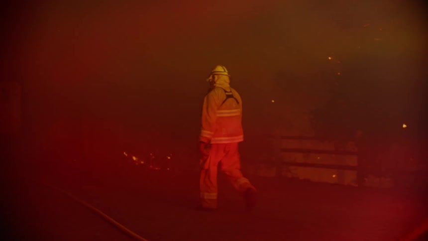 A firefighter walks through heavy smoke toward a burning forest in the distance