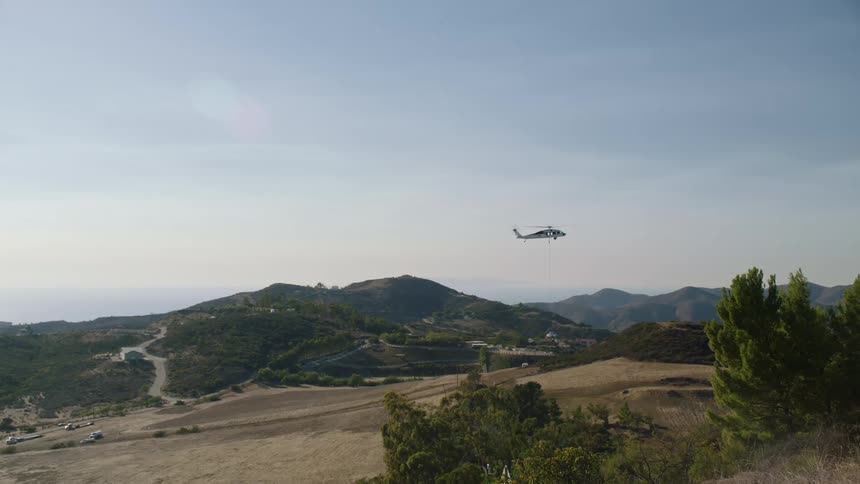A helicopter is landing on an empty plain before some distant forests and hill peaks
