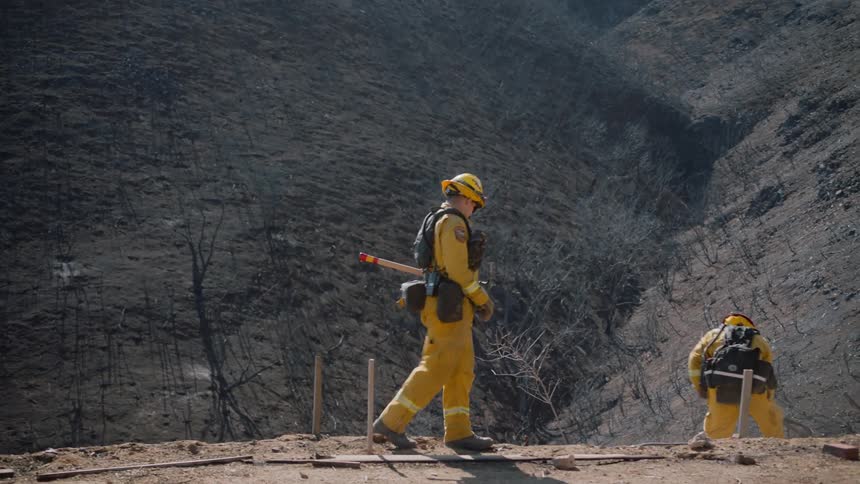 One firefighter walks toward where another firefighter is digging at smoking ground near edge of burnt mountain slopes
