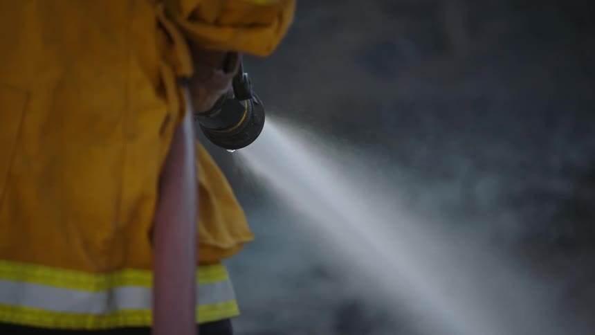 A firefighter holds a hose as water flies out and there is smoke in the distance