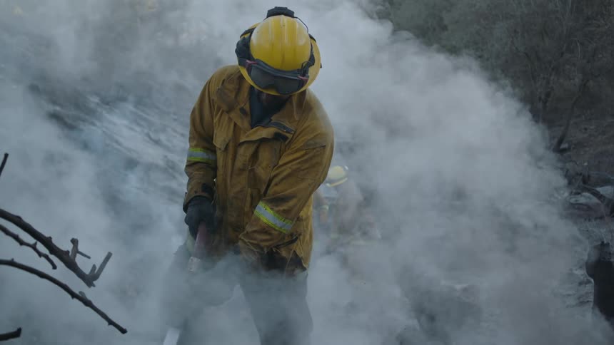 A firefighter is standing outside holding a hose as smoke fills the air around him
