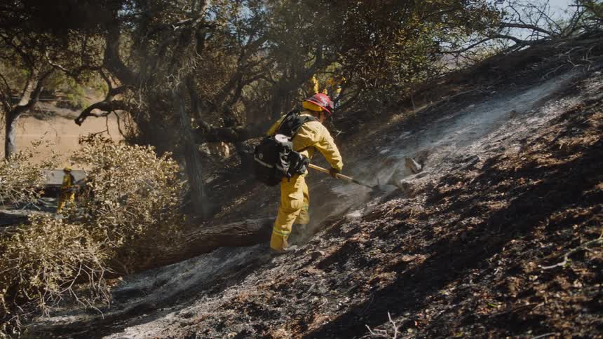 Firefighter digs at ground on slope covered in ash while other firefighters move in background