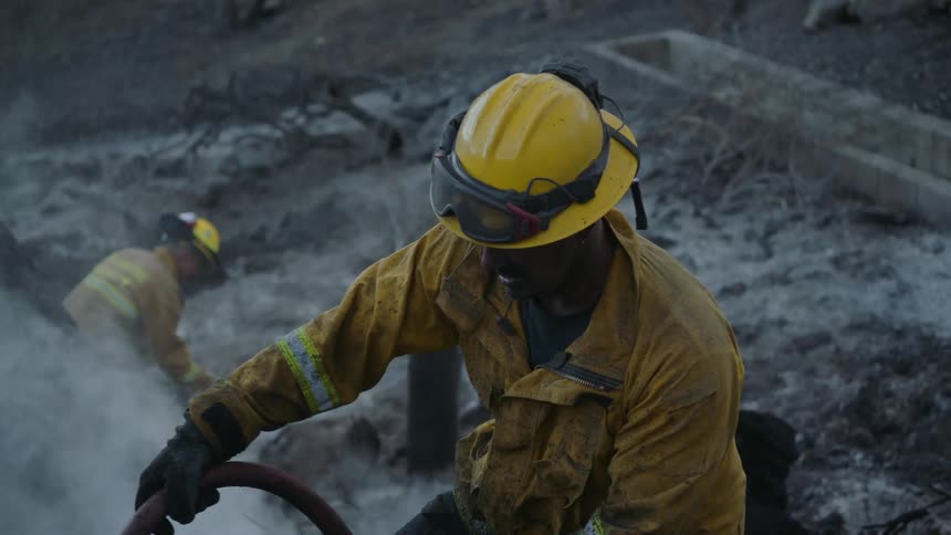 A firefighter walking with a hose and turning it off then walking forward while two other firemen work in the background