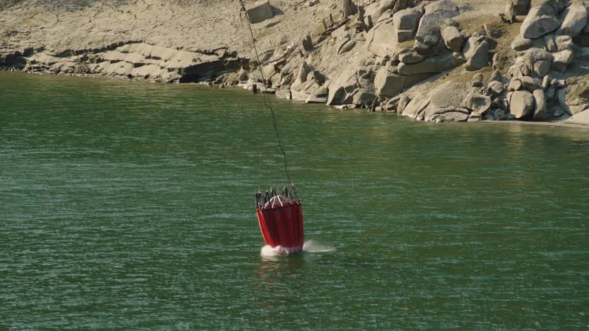 Red helicopter bucket splashes into green water near boulder strewn shore