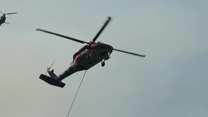 A helicopter carries a specialized bucked suspended on a cable to deliver water for aerial firefighting