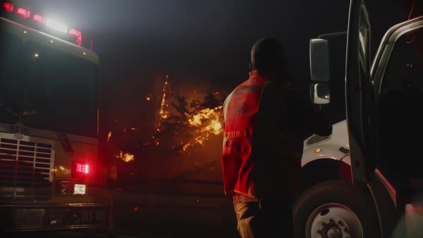 Firefighters near vehicles looking a wildfires burning trees on a hill at night