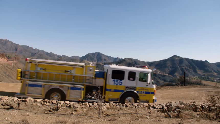A yellow fire truck drives across an empty dirt plateau in front of brown cliff peaks