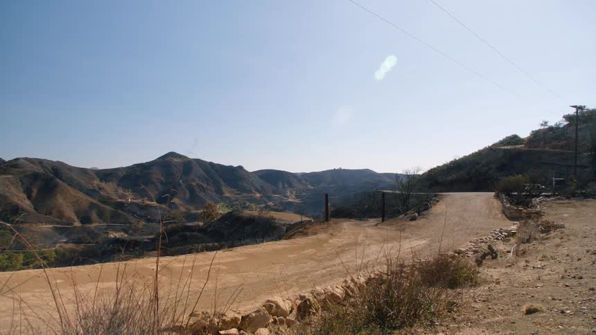 The sun shines on a dirt road in a rural area with mountains in the distance under a clear sky
