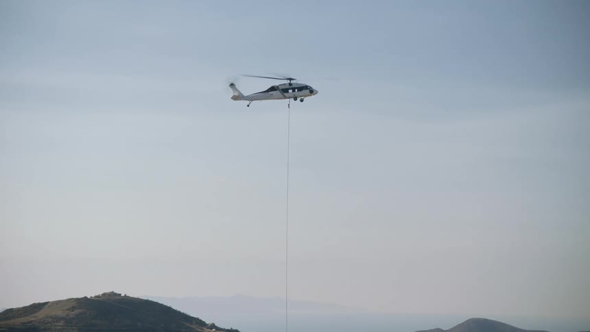 A helicopter carries a specialized bucked suspended on a cable to deliver water for aerial firefighting